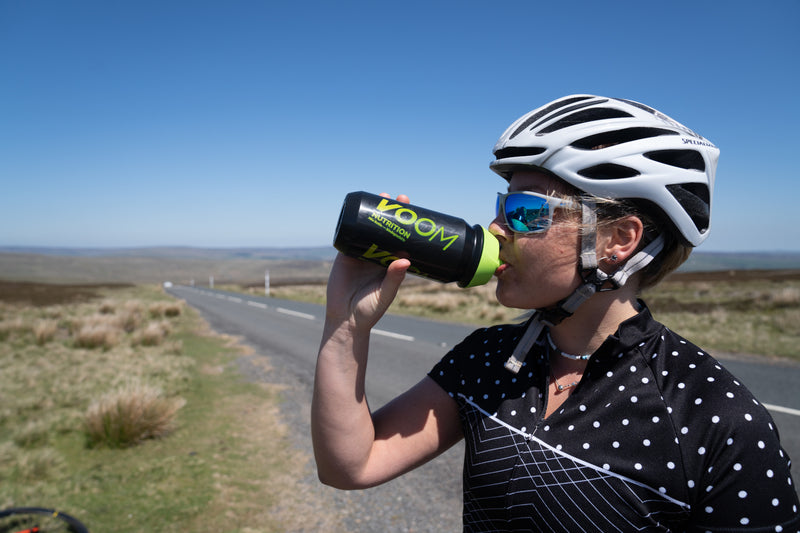 A female cyclist stood beside the road to drink from a black and green VOOM Nutrition biodegradable sports water bottle.
