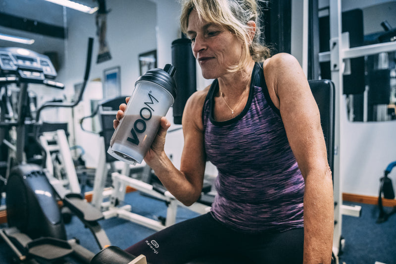 A lady sits on a weights machine in a gym drinking VOOM Chocolate protein recovery shake after a gym workout.