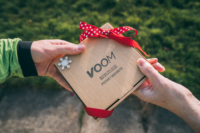 A Voom branded brown cardboard box with a red ribbon being passed from one hand to another hand