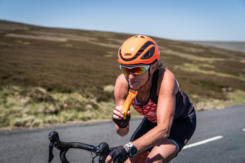 A female cyclist rides up a steady hill, one hand on the handlebars and the other holding a VOOM Beta Blast pocket rocket, about to take a bite